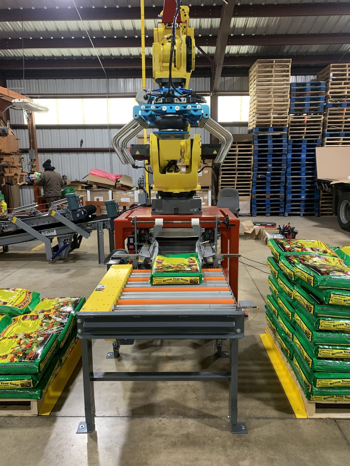 A FANUC palletizing robot lifting a bag from the infeed conveyor, with built pallets of product either side of the cell.