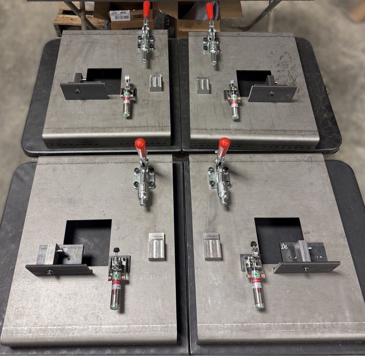Four matching weld fixtures built by TMD, laid out two by two — each a steel base plate with machined locating blocks, a red-handled toggle clamp and a pneumatic clamp cylinder.